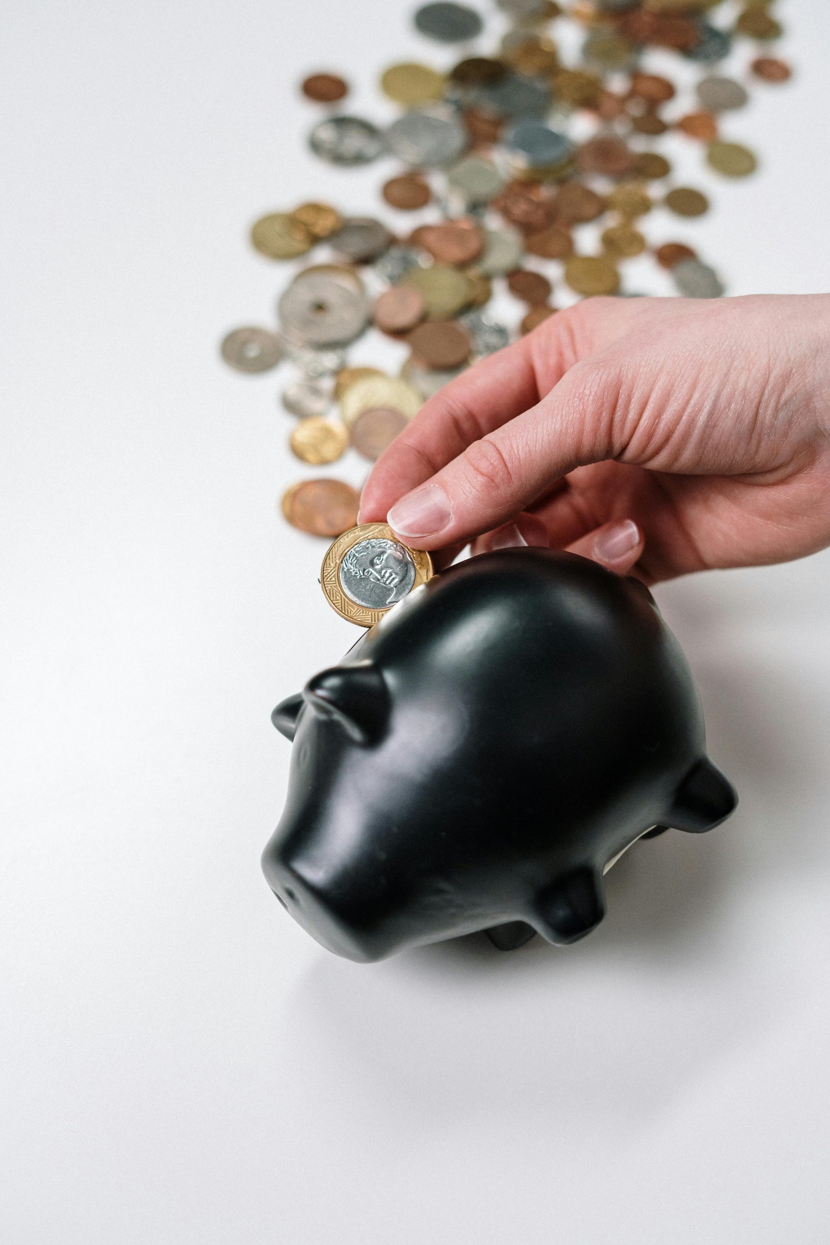 A hand putting a coin into a black piggy bank with scattered coins on a white background.