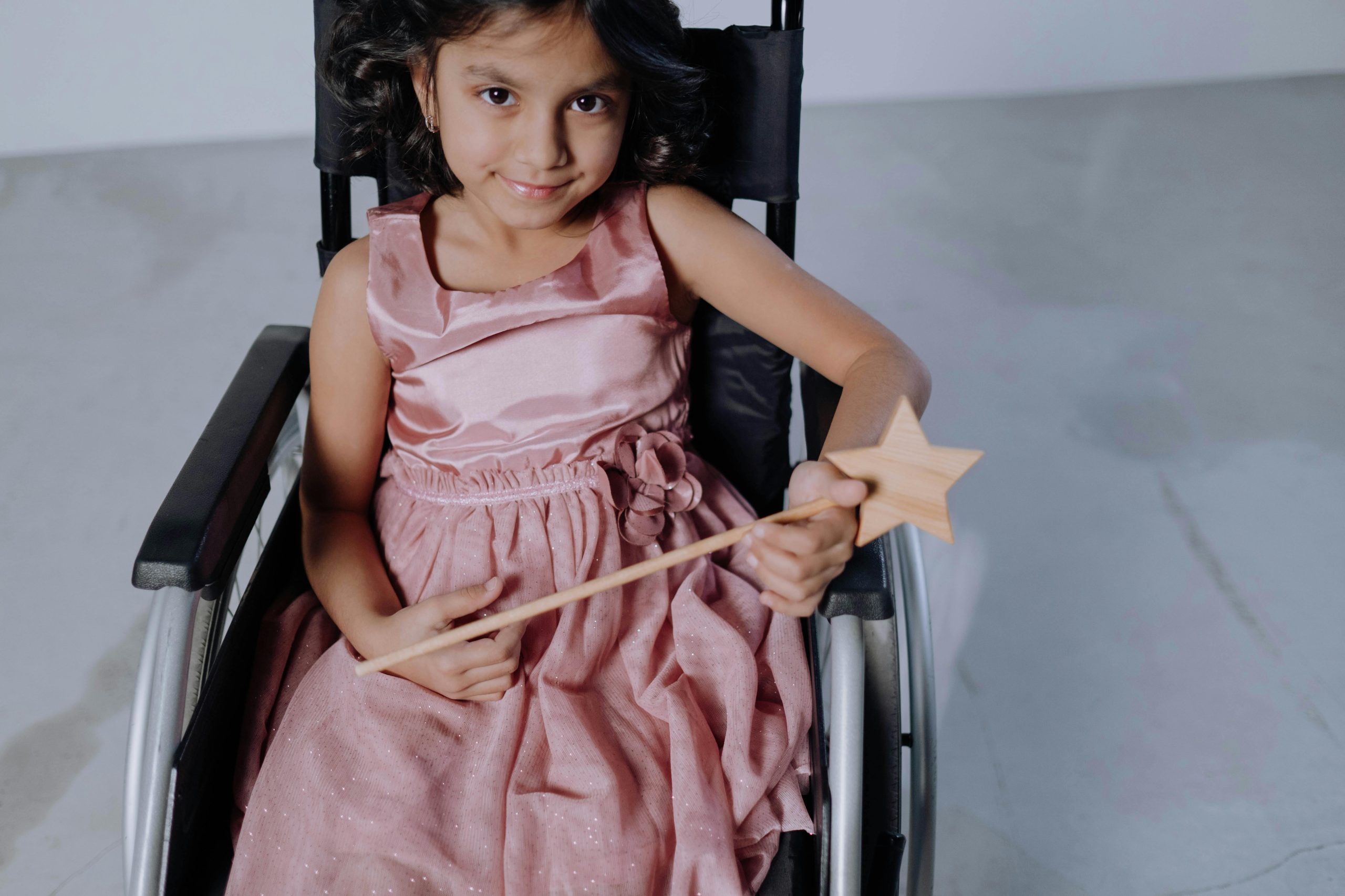 Charming young girl in wheelchair joyfully holding a wooden magic wand indoors.
